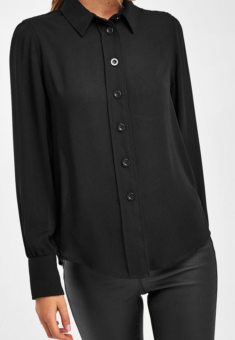 Chemise noire à manches longues en tissu lisse, avec ouverture boutonnée sur le devant, col classique et détails légèrement froncés aux épaules.