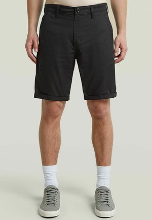 Homme portant un short noir retroussé, des chaussettes blanches mi-mollet et des baskets grises à lacets, debout devant un fond uni.