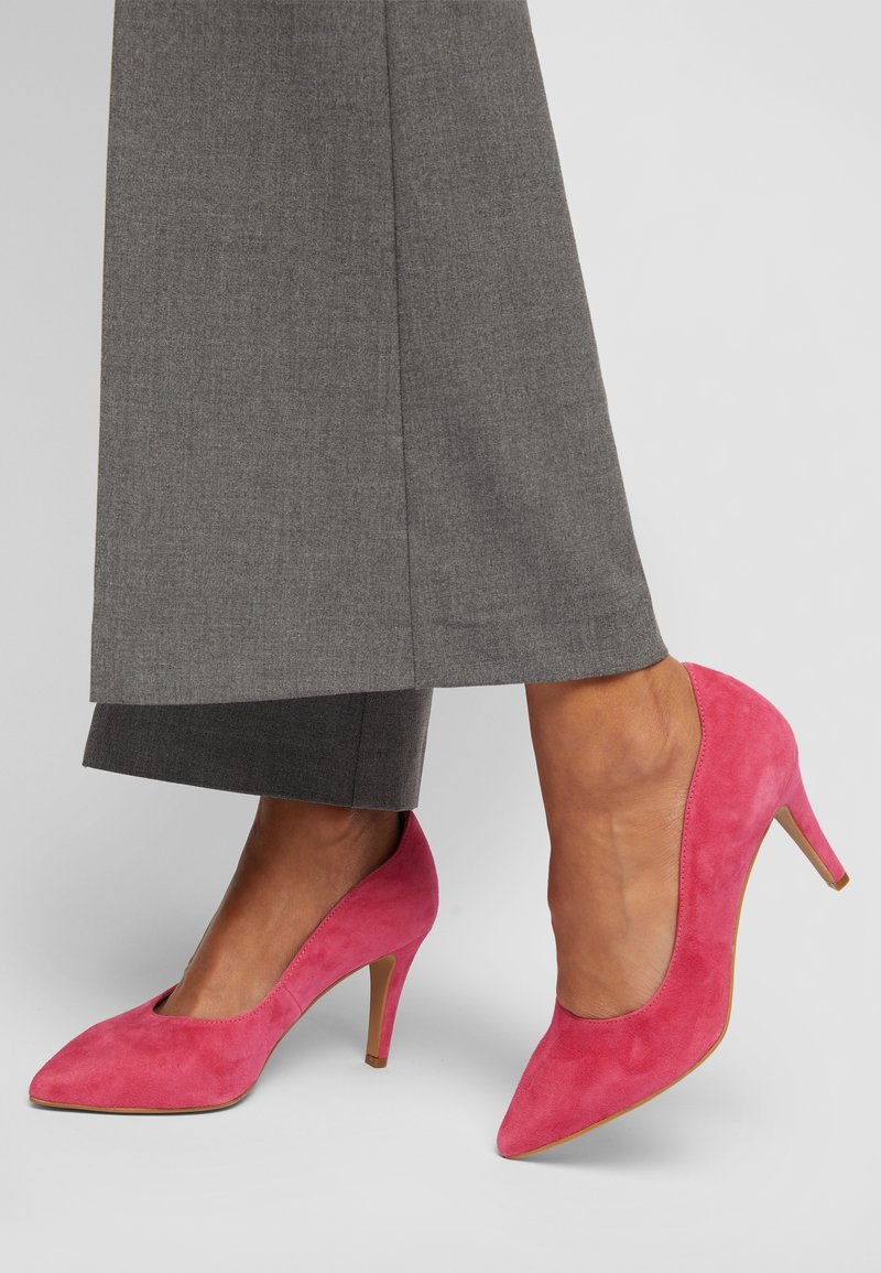 Pieds portant des escarpins en daim rose vif associés à un pantalon gris à jambes larges sur un fond uni.