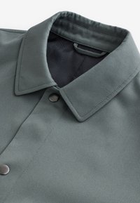 Gros plan d'une veste col vert-gris avec une doublure intérieure noire texturée et deux boutons-pression métalliques visibles.