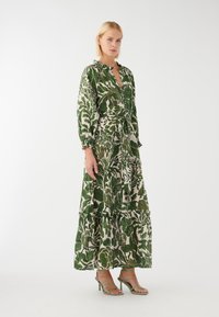 Grünes, blumig gemustertes Kleid mit langen Ärmeln, Rüschensaum, Taillengürtel und gestuftem Saum. Kombiniert mit grünen Riemchenschuhen.