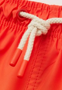 Los pantalones de baño rojos cuentan con una cintura texturizada con un cordón trenzado blanco, que termina en puntas de plástico rojas, y detalles de costura visibles.