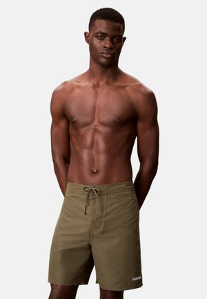 Uomo muscoloso in piedi con le mani dietro la schiena, indossa pantaloncini da bagno Calvin Klein verde oliva su uno sfondo chiaro e uniforme.
