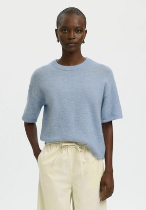 Kvinde med kort hår iført lyseblå kortærmet sweater og cremefarvede bukser med snøre, står med den ene hånd i lommen.