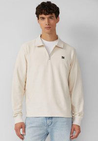 Hellbeiger Pullover mit Reißverschlusskragen und gerippter Textur. Ausgestattet mit einem kleinen Logo auf der Brust, langen Ärmeln und einem lockeren Schnitt.