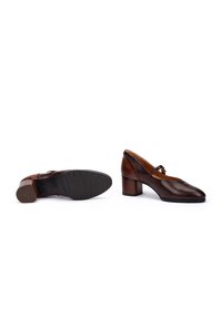 Chaussures à talons en cuir marron avec un bout rond et un design à sangles croisées. Talon texturé et semelle en caoutchouc avec des rainures pour une meilleure adhérence.