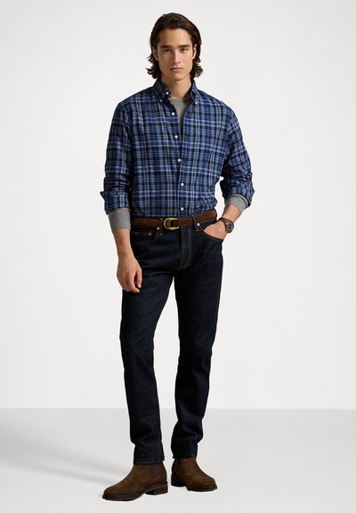 Camicia a quadri blu sopra un maglione grigio, jeans scuri e stivaletti in suede marrone, con una cintura marrone e un orologio da polso.