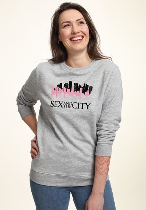 Uśmiechnięta kobieta o brązowych włosach ubrana w szarą bluzę "Seks w wielkim mieście", z czerwonym tatuażem z henny na lewym przedramieniu, pozuje z ręką na biodrze.