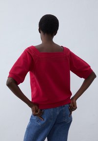Maglione rosso con scollatura quadrata, maniche corte e dettagli di cucitura decorativa lungo il corpo; indossato con jeans in denim blu.