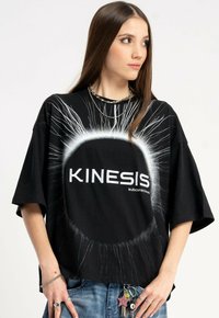 Młoda kobieta ubrana w czarną, dużą koszulkę z napisem "KINESIS subconscious" oraz białym promieniującym wzorem w kształcie koła, zestawioną z dżinsami i dodatkami.