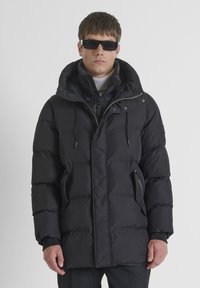 Giubbotto imbottito nero con colletto alto, zip frontale e cappuccio regolabile con cordino. Presenta tasche laterali con zip e texture trapuntata.