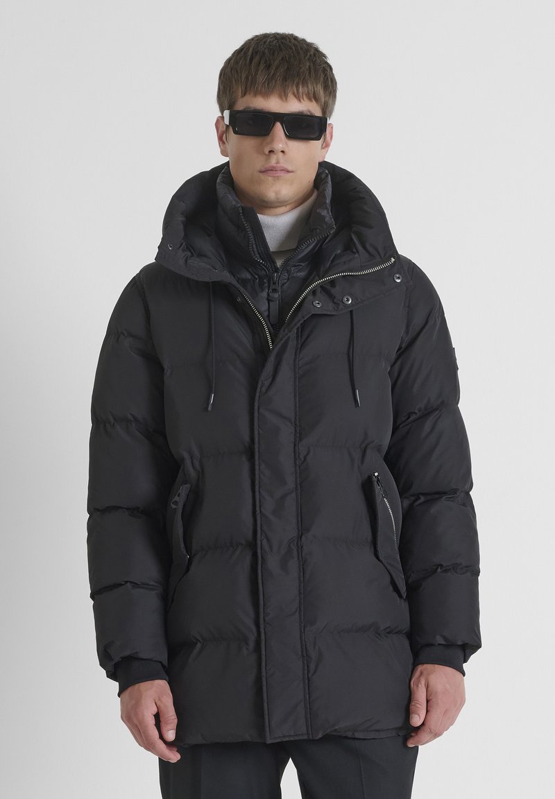 Giubbotto imbottito nero con colletto alto, zip frontale e cappuccio regolabile con cordino. Presenta tasche laterali con zip e texture trapuntata.