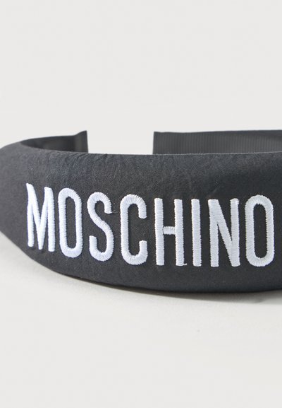 Μαύρη υφασμάτινη κορδέλα με λευκή κεντημένη επιγραφή "MOSCHINO" στο μπροστινό μέρος, τοποθετημένη πάνω σε ένα απλό ανοιχτό φόντο.