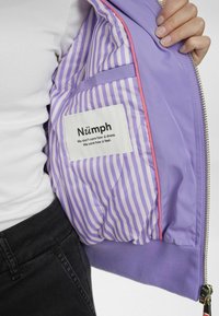 Violeta bomber jakas iekšpuse ar lavandas un baltām svītrām, rozā maliņa, etiķete ar zīmola nosaukumu un quiltēta tekstūra.