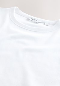 Gros plan d’un t-shirt blanc côtelé à col rond avec une étiquette de marque visible "NEXT" au niveau du col.