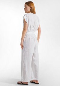 Mujer con cabello rojo de perfil, vestida con un mono blanco sin mangas y sandalias negras, sobre un fondo blanco liso.