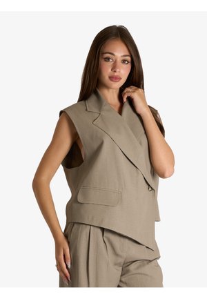 Giovane donna con lunghi capelli castani indossa una giacca beige senza maniche e pantaloni coordinati, posando con una mano vicino al viso e l'altra sul fianco.