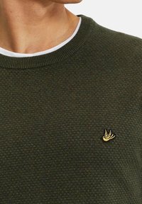 Tmavě zelený pletený svetr s texturovaným vzorem, kulatým výstřihem a malým žlutým logem ptáčka na levé straně hrudi.