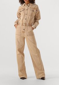 Vrouw in een beige jumpsuit met knopen, voorzien van borst- en zijzakken, elastische taille, staand met handen in de zakken en bruine schoenen.