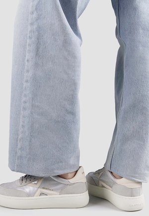 Person trägt hellblaue Jeans mit weitem Bein und hellgraue Sneakers mit dicken weißen Sohlen und steht mit leicht gespreizten Füßen.