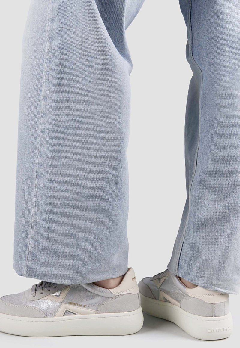 Personne portant un jean bleu clair à jambes larges et des baskets gris clair avec des semelles épaisses blanches, debout les pieds légèrement écartés.