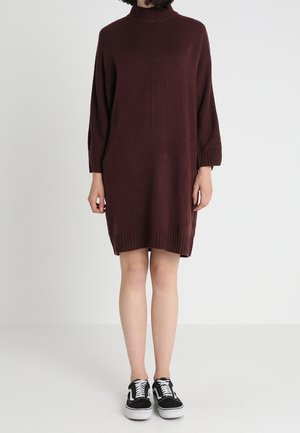 Kvinde iført en mørkerød strikket langærmet kjole og sort-hvide lave sneakers, der står på en ensfarvet lys baggrund.