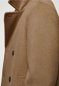 Abrigo de lana marrón con cuello y dos botones negros, con una textura suave y un corte ajustado. Detalle de bolsillo lateral visible.