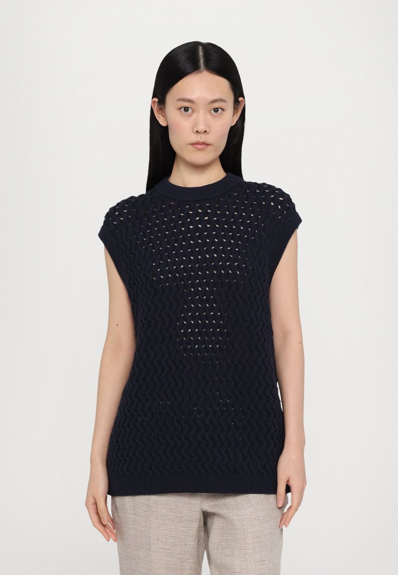 Mornarsko plavi pleteni bez rukava s otvorenim uzorkom i rebrastim rubovima na izrezu i rubu. Uparen s svijetlim bež hlačama.