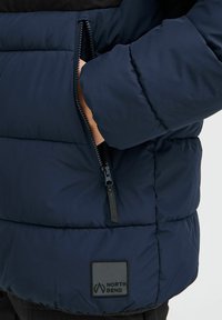 Mørkeblå pufferjakke med lynlåslomme, der har en sort øverste del og et sort logo-patch med teksten "NORTH BEND." Vatteret tekstur.