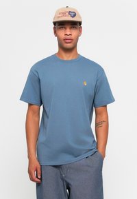 Homme portant une casquette beige avec du texte, une chemise bleu à manches courtes avec un petit logo sur la poitrine, et un pantalon gris ample, se tenant devant un fond blanc.