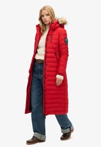 Rød polstret lang jakke med hætte med pelskant, lynlås og trykknaplukninger samt et logo patch på ærmet. Bæres over en beige sweater og brede jeans.