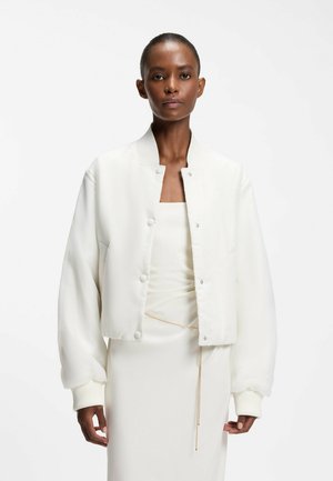 Femme aux cheveux courts portant une veste bomber blanche rembourrée sur une robe blanche avec une fine ceinture chaîne dorée sur un fond blanc uni.