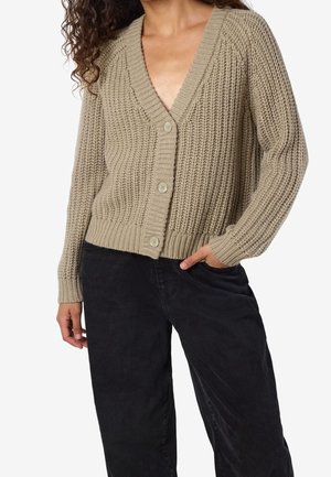 Femme portant un cardigan beige en grosse maille boutonné et un pantalon noir ample, main dans la poche, sur fond blanc.