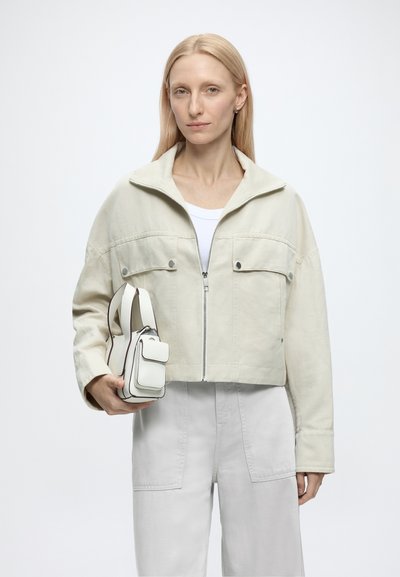 Femme aux cheveux blonds lisses portant une veste et un pantalon beige clair, tenant un petit sac à main blanc sur un fond clair uni.