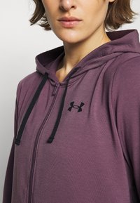 Fialová mikina na zip s černým logem na prsou, která má kapuci se stahovací šňůrkou a texturovanou látku.