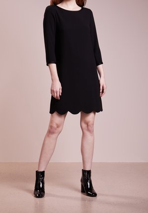 Žena v černých šatech po kolena s vroubkovaným lemem a tříčtvrtečními rukávy, doplněná černými lesklými kotníkovými botami.