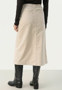 Fustă din denim bej, de lungime medie, cu o textură simplă și buzunare la spate, purtată cu cizme negre din piele lungi.