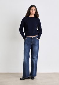 Suéter corto de punto acanalado navy, combinado con jeans de mezclilla azul oscuro de pierna recta. Zapatos negros, manos en los bolsillos, fondo neutro.