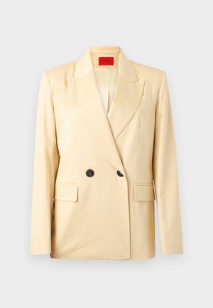 Μπεζ διπλόπινο σακάκι με φαρδιές πέτρες, δύο μαύρα κουμπιά, τσέπες με καπάκια και κόκκινη εσωτερική ετικέτα σε ανοιχτό φόντο.