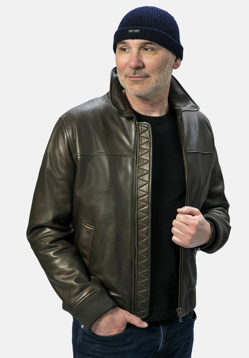 Braune Lederjacke mit Reißverschluss vorne, gerippten Bündchen und dekorativer Naht entlang der Vorderseite. Getragen mit einem schwarzen Hemd und einer marineblauen Mütze.