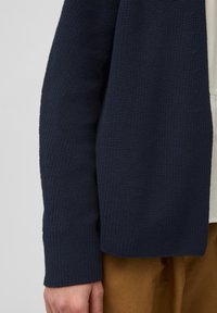 Granatowy dzianinowy kardigan o teksturowanej tkaninie, z luźnym krojem i szerokimi rękawami, zestawiony z jasnym topem i brązowymi spodniami.