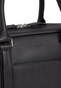 Bolso de cuero negro texturizado con dos asas superiores, cremallera metálica y un sutil grabado del logo. Presenta líneas limpias y una forma rectangular.