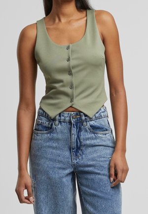 Femme portant un haut olive sans manches à boutons et un jean en denim bleu taille haute, debout devant un fond gris.