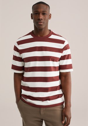 Homme debout les mains dans les poches, portant une chemise à manches courtes à rayures horizontales blanches et rouge foncé, et un pantalon beige, sur un fond uni.