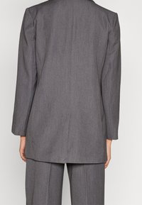 Blazer grigio su misura con taglio dritto, maniche lunghe e una texture di tessuto liscia. Dettaglio della cucitura sul retro ben visibile.