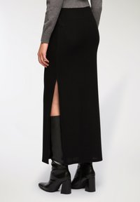 Gonna maxi nera a costine con spacco laterale, realizzata in un tessuto testurizzato. Abbinata a stivali lucidi neri con tacco al ginocchio.