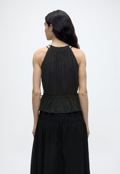 Γυναίκα με μακριά σκούρα μαλλιά που φοράει μαύρο αμάνικο τοπ με halter και μαύρη φούστα, βλέποντας από πίσω με φόντο μονόχρωμο.