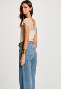 Beige top med blondedetalje på ryggen, parret med lyseblå højtaljede jeans. Guld accents armbånd og bælte inkluderet.