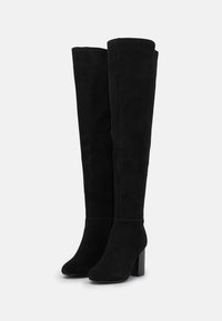 Pár černých semišových vysokých kozaček s blokovými podpatky a bočními zipy, mírně nakloněné doleva na bílém pozadí.