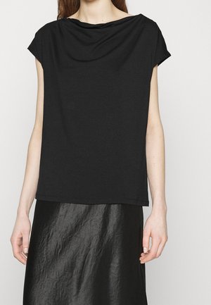 Kobieta ubrana w czarny top bez rękawów z drapowanym dekoltem i czarną teksturowaną spódnicę na jednolitym tle.